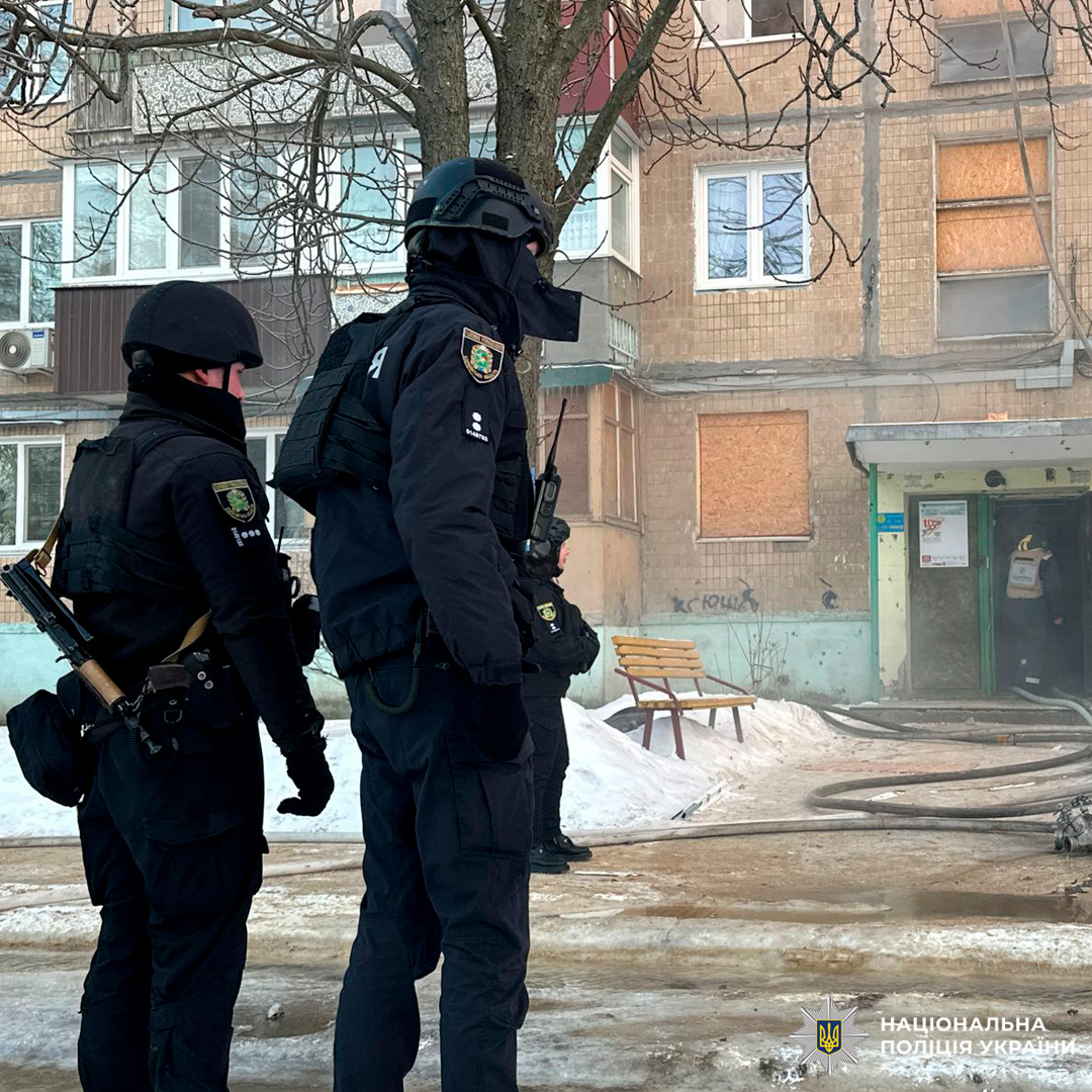 Група поліцейських у шоломах та бронежилетах забезпечує безпеку та перевіряє пошкоджений будинок після надзвичайної події, на тлі зимових умов.