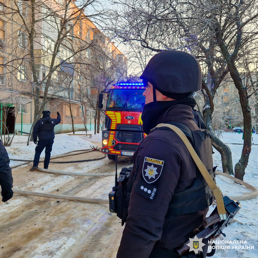 Пожежна машина з увімкненими проблисковими маячками, розгорнуті рукави для гасіння, поліцейські у спорядженні координують дії біля багатоповерхового будинку.
