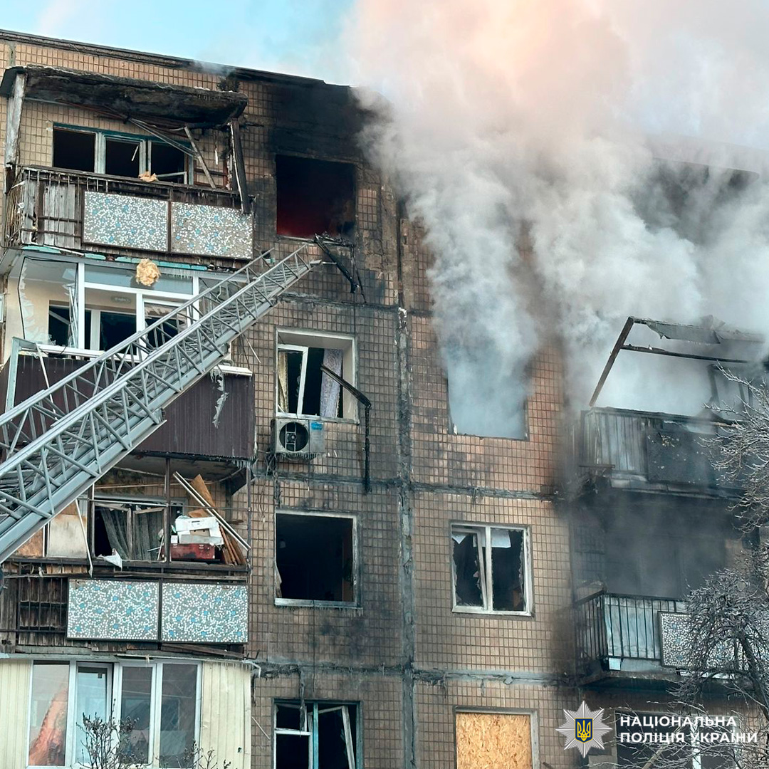 Багатоповерховий житловий будинок із пошкодженими фасадом та балконами, густий дим із верхніх поверхів, пожежна драбина піднята для ліквідації наслідків.