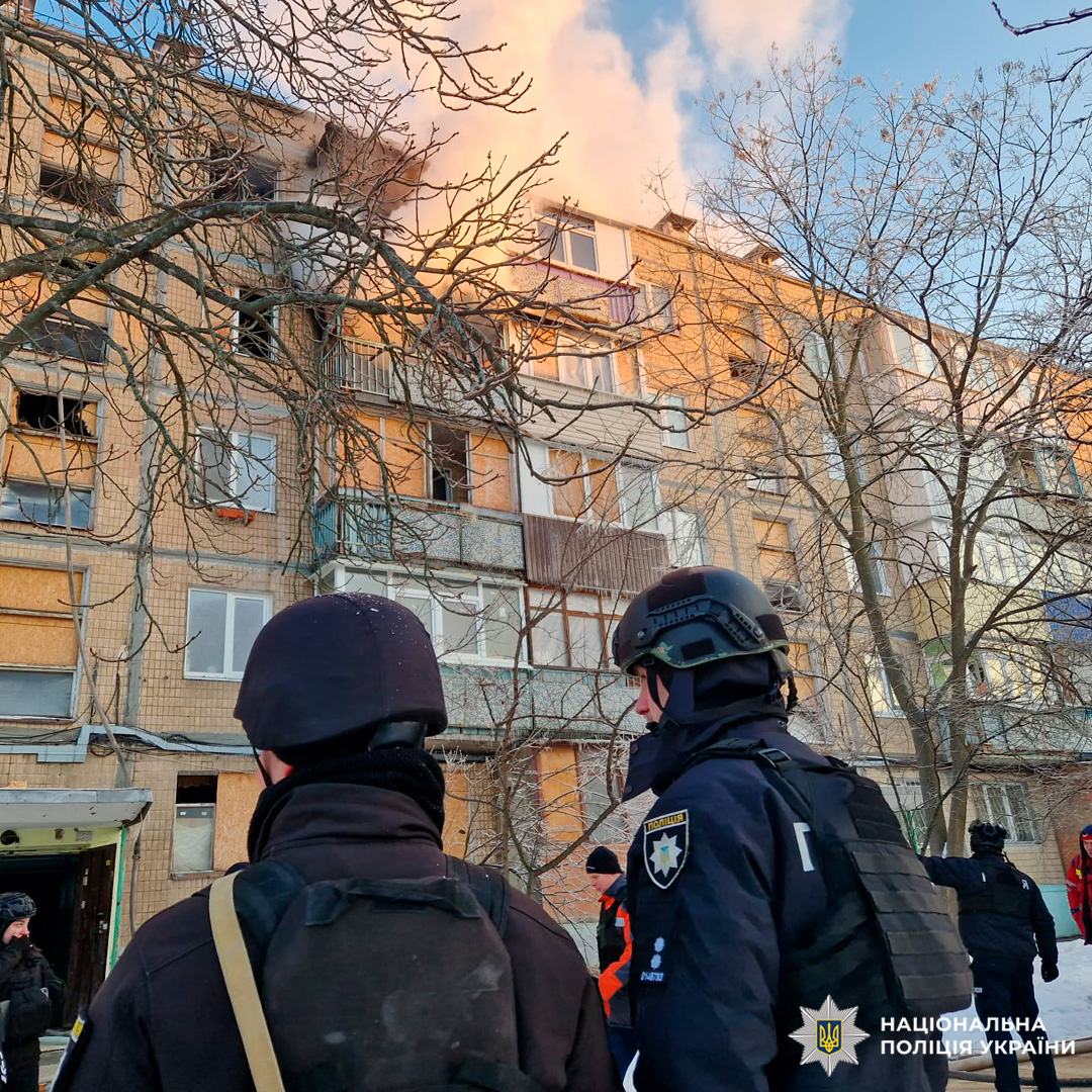 Поліцейські України перед пошкодженим житловим будинком, з якого йде дим, зимова пора.