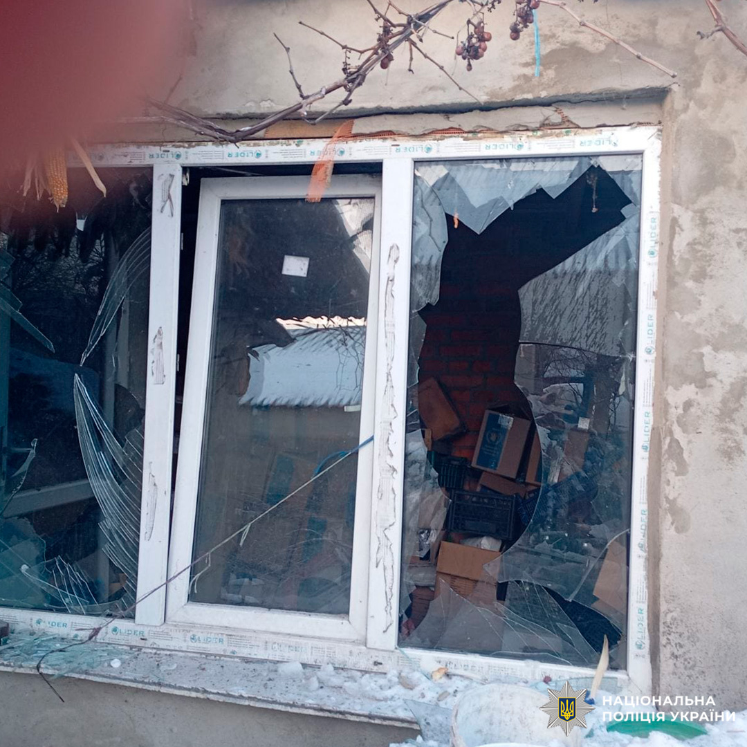 Розбите вікно з тріщинами та великими отворами у склі, всередині видно коробки й цегляну стіну, зовні сніг та сухі виноградні грона.