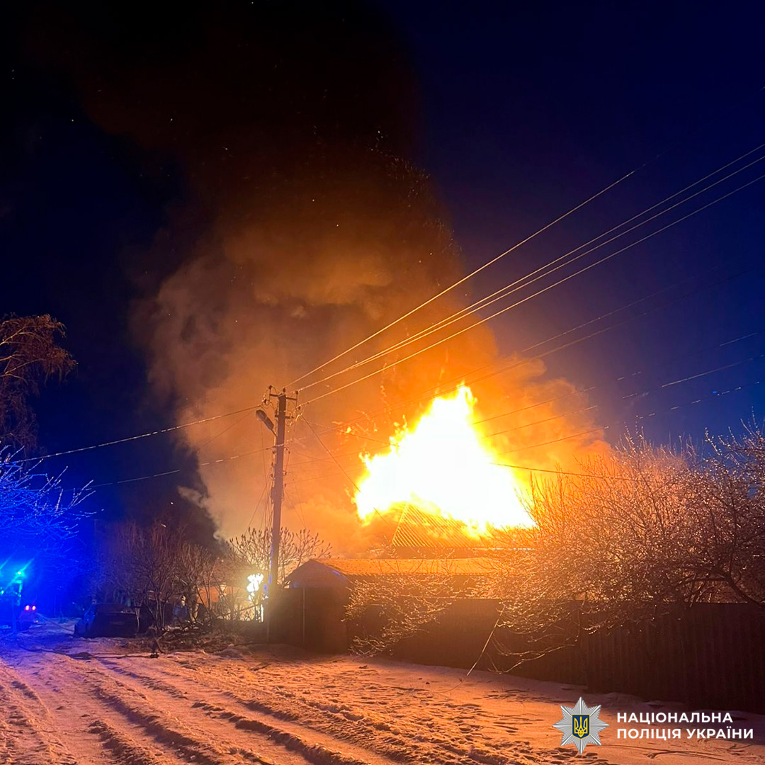 Велике займання даху будівлі в нічний час, яскраве полум’я освітлює околиці, густий дим здіймається в небо, на землі та деревах лежить сніг, видно електродроти й синє світло спецтехніки.