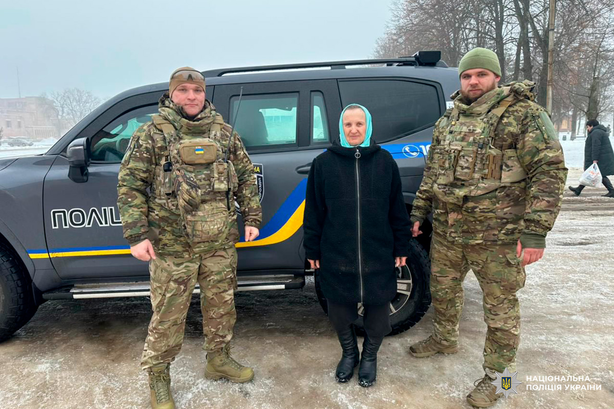 Двоє військових у камуфляжній формі стоять по обидва боки від літньої жінки в чорному пальті та світлій хустці, позаду темний поліцейський автомобіль.