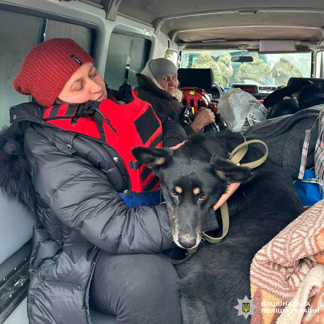 Усередині транспортного засобу двоє осіб у теплих речах, один у червоній шапці тримає великого чорного собаку з рудими відмітинами, позаду багаж і речі.