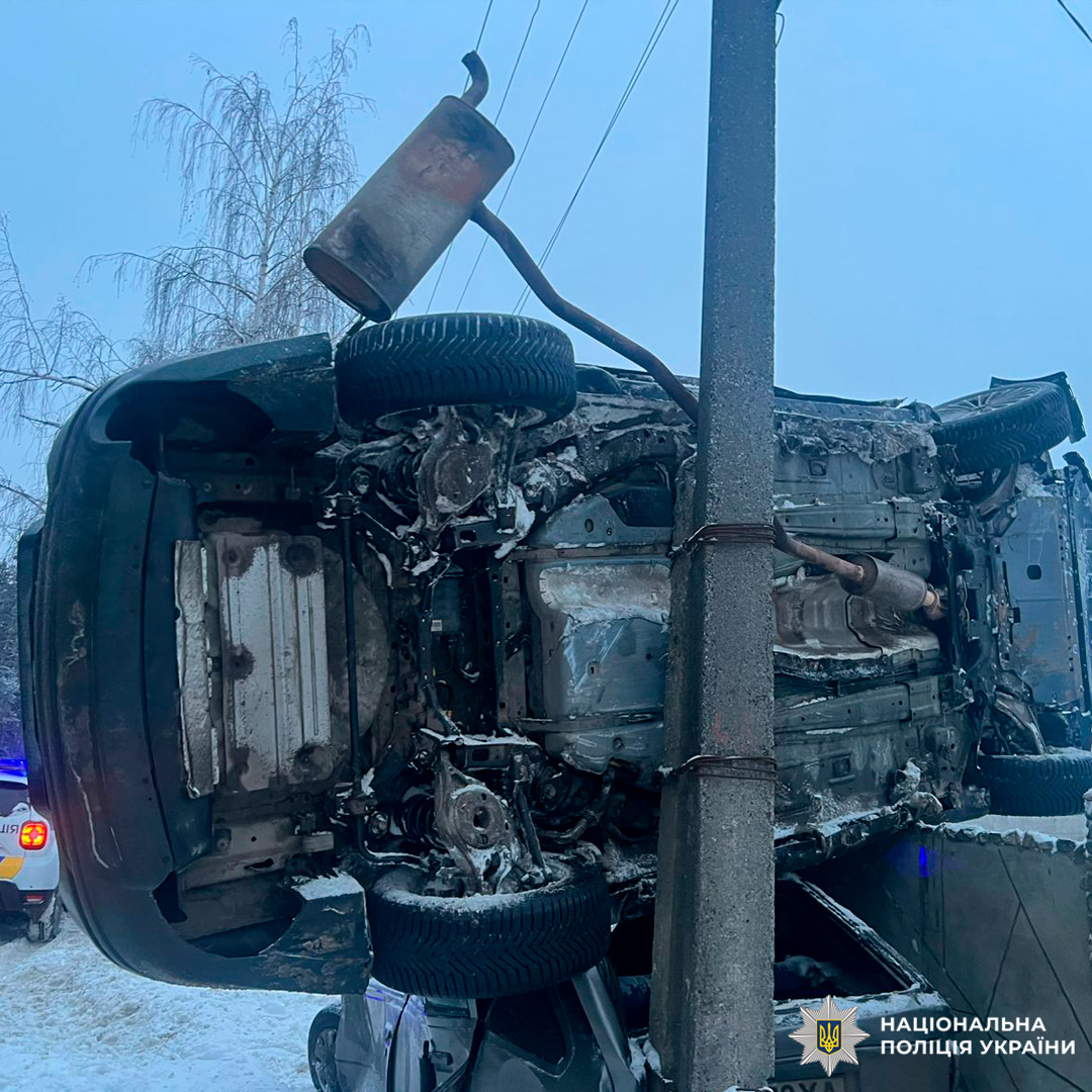 Автомобіль після зіткнення вертикально притиснутий до електроопори, видно пошкодження та від’єднані деталі, поруч службовий транспорт із увімкненими маячками.