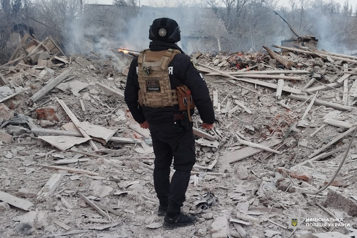 Співробітник Національної поліції України оглядає місце руйнувань після вибуху, серед уламків та диму.