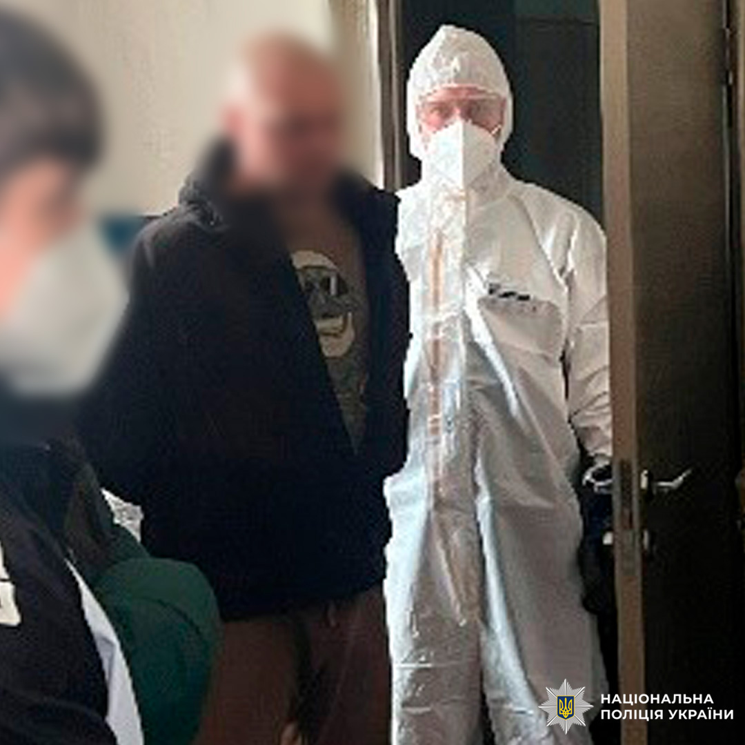 Працівник поліції у захисному костюмі проводить операційні дії в приміщенні, поруч цивільні особи.