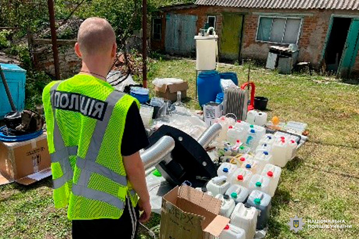 Поліцейський контролює вилучені каністри, бочки та обладнання, розкладені на подвір’ї під час операції.