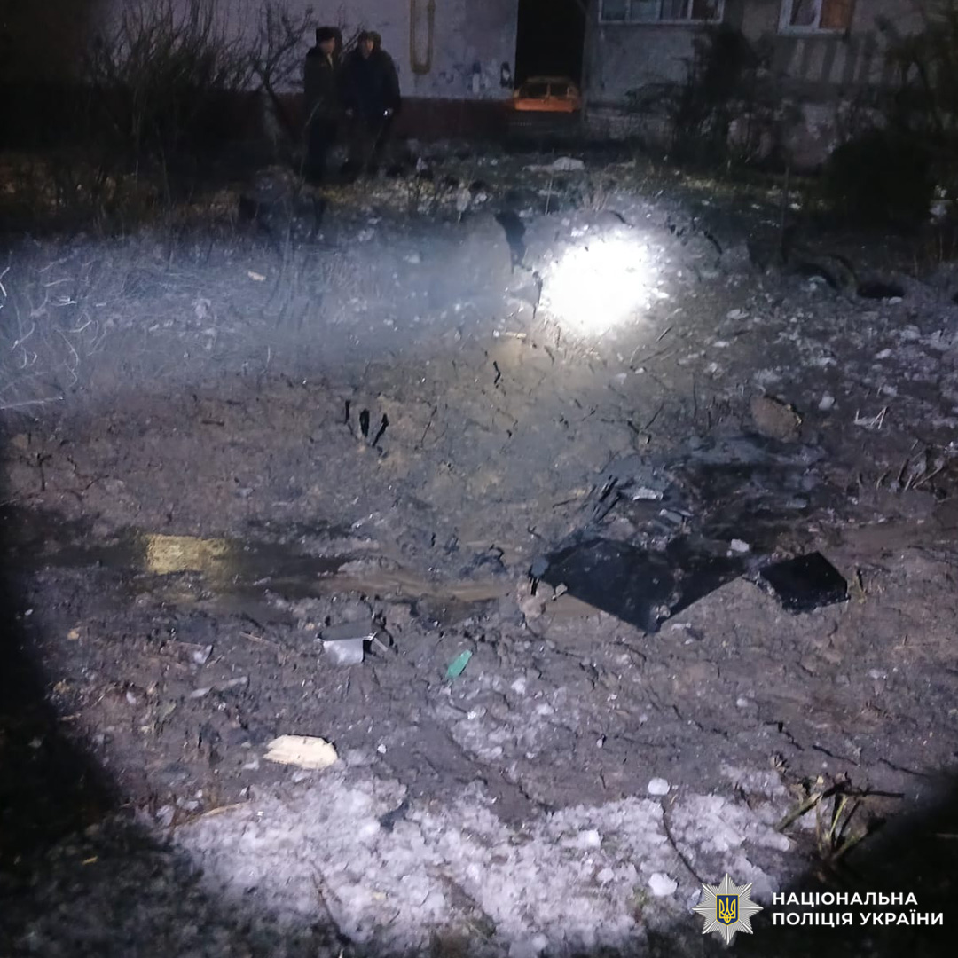 Кратер від вибуху в житловому районі, навколо — уламки та пошкоджений ґрунт. На задньому плані видно людей біля пошкодженої будівлі.