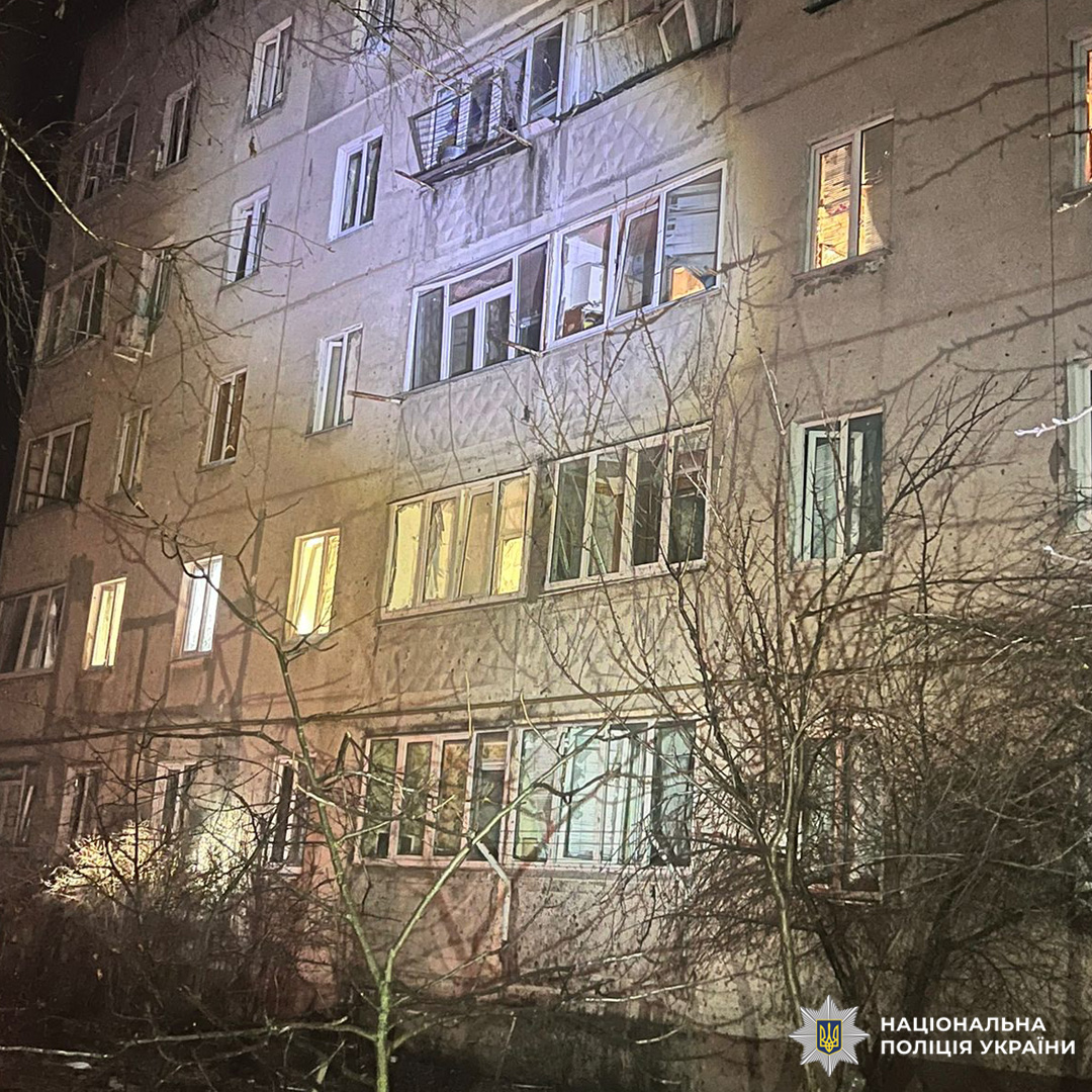 Нічний вигляд багатоповерхового житлового будинку з освітленими вікнами, на передньому плані - дерева без листя.