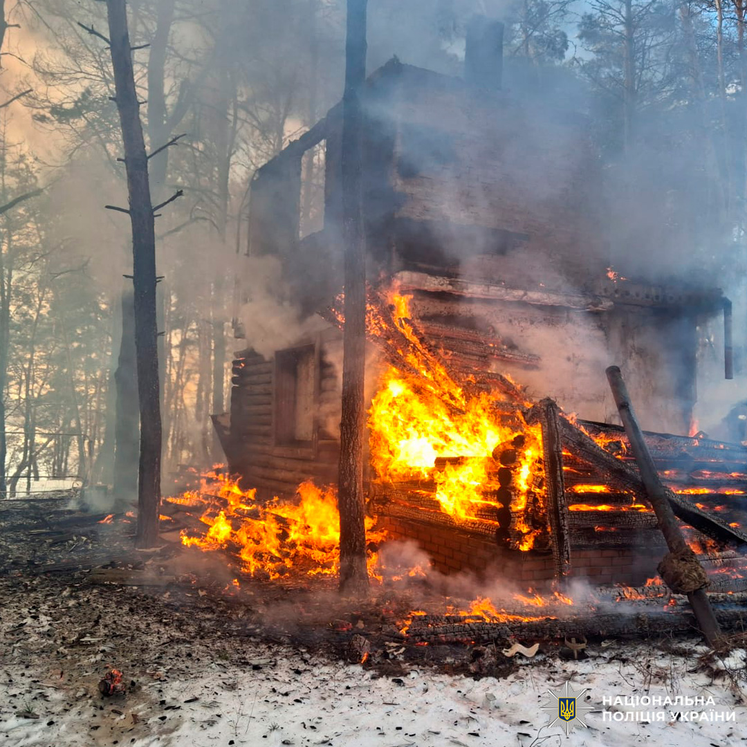 Дерев’яний будинок у лісистій місцевості повністю охоплений вогнем, густий дим піднімається над дахом.