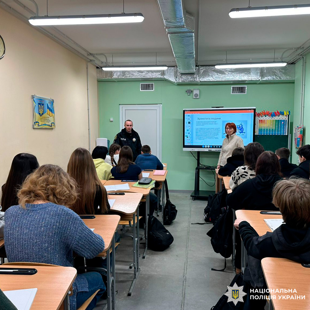 Учні сидять у класі, слухаючи виступ поліцейського та цивільного представника перед дошкою й монітором.