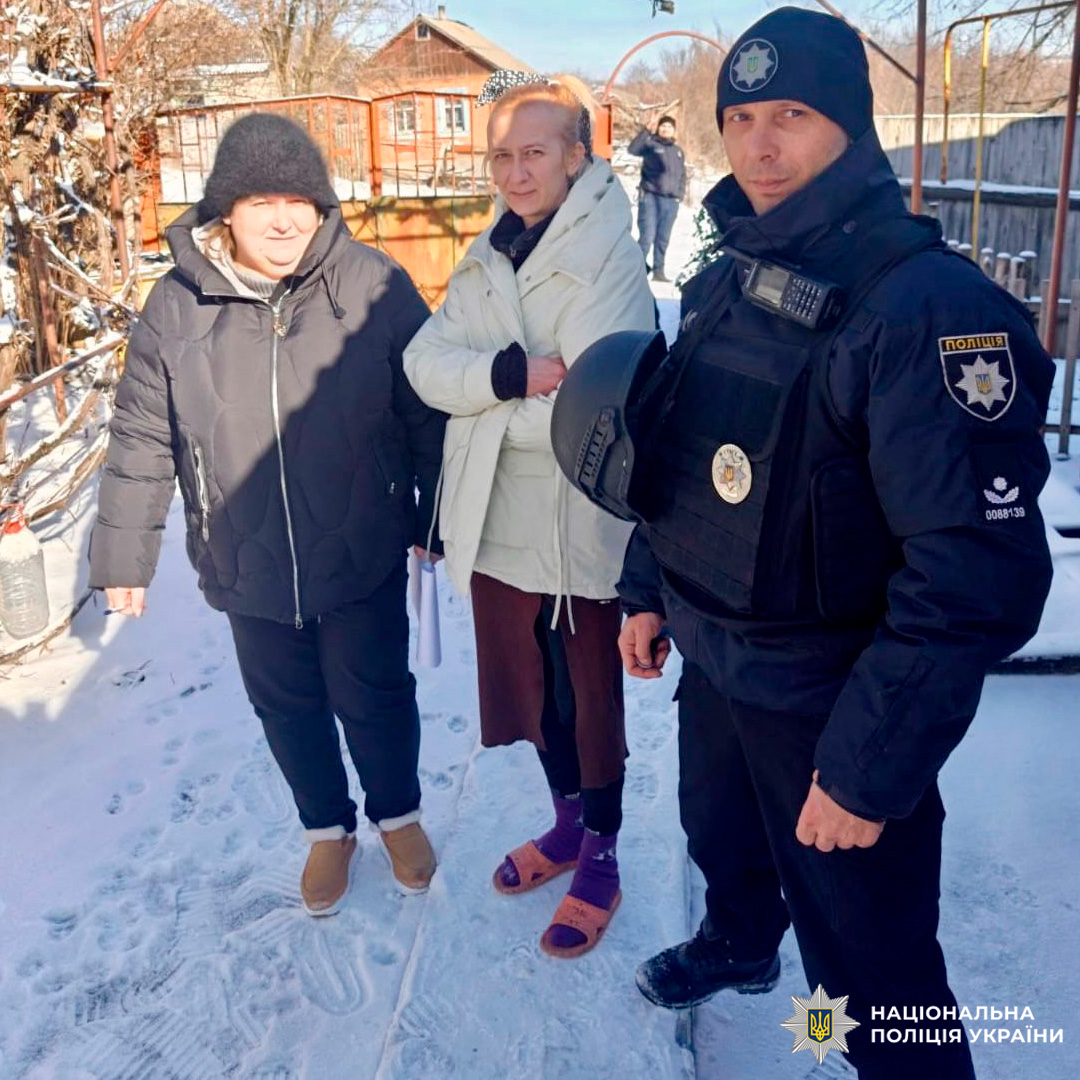 Поліцейський у формі з тактичним жилетом і шоломом стоїть на засніженій вулиці поруч із двома людьми в зимовому одязі.