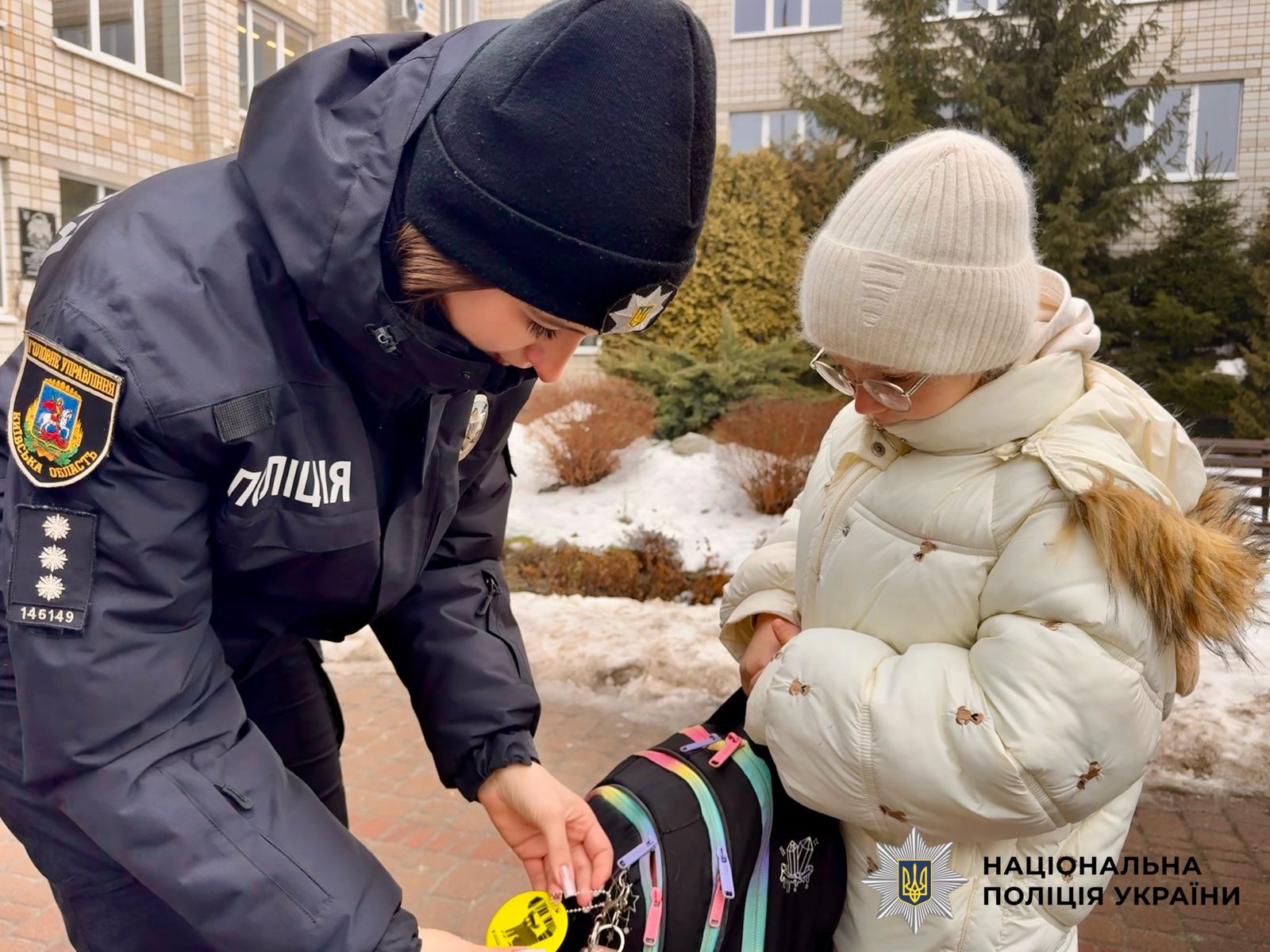 На фотографії зображена поліцейська Київщини, яка допомагає дитині чіпляти світлоповертальний флікер на рюкзак.