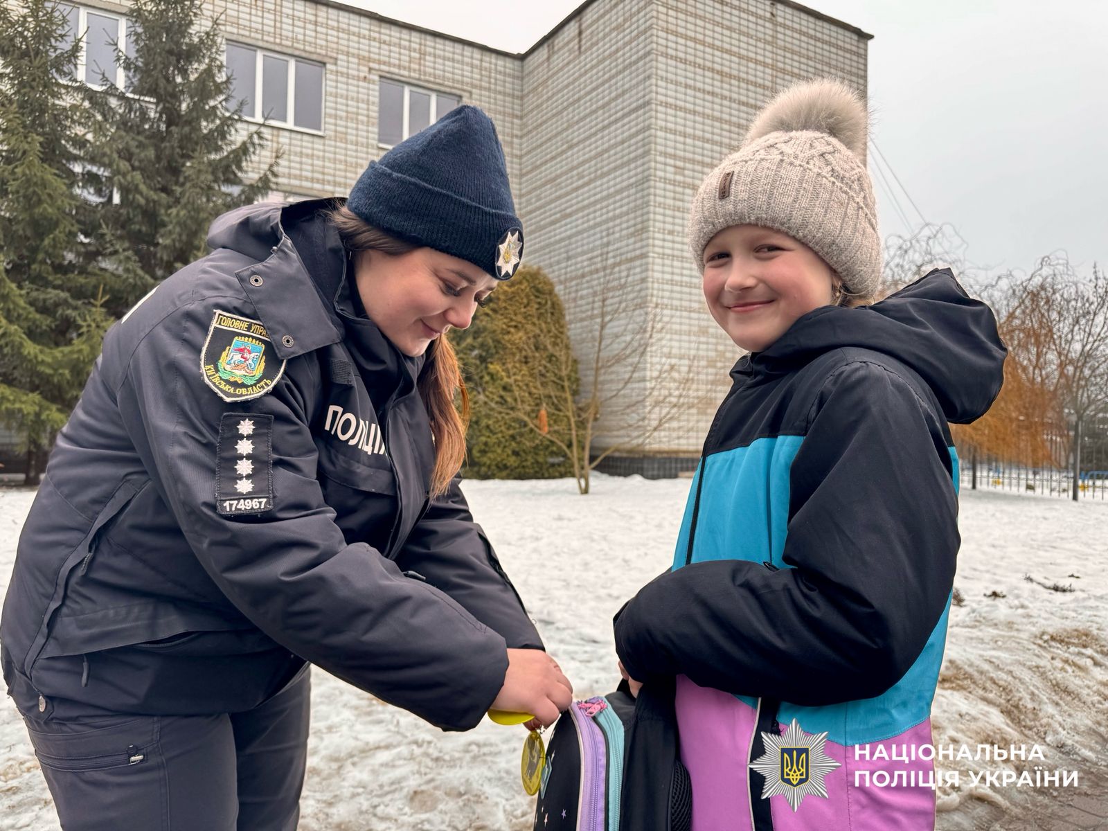 На фотографії зображена поліцейська Київщини, яка допомагає дитині чіпляти світлоповертальний флікер на рюкзак.