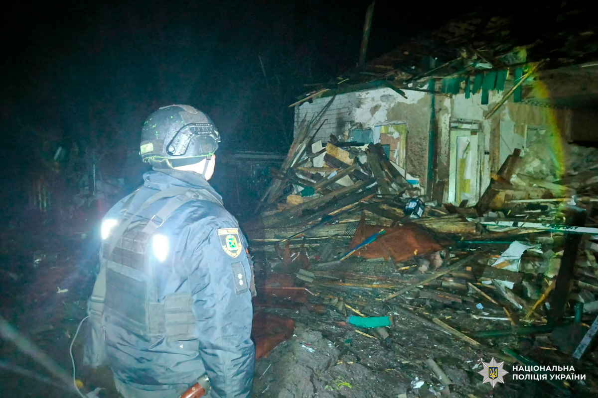 Поліцейський у бронежилеті та касці проводить огляд будівлі з відкритими дверима та слідами руйнувань.