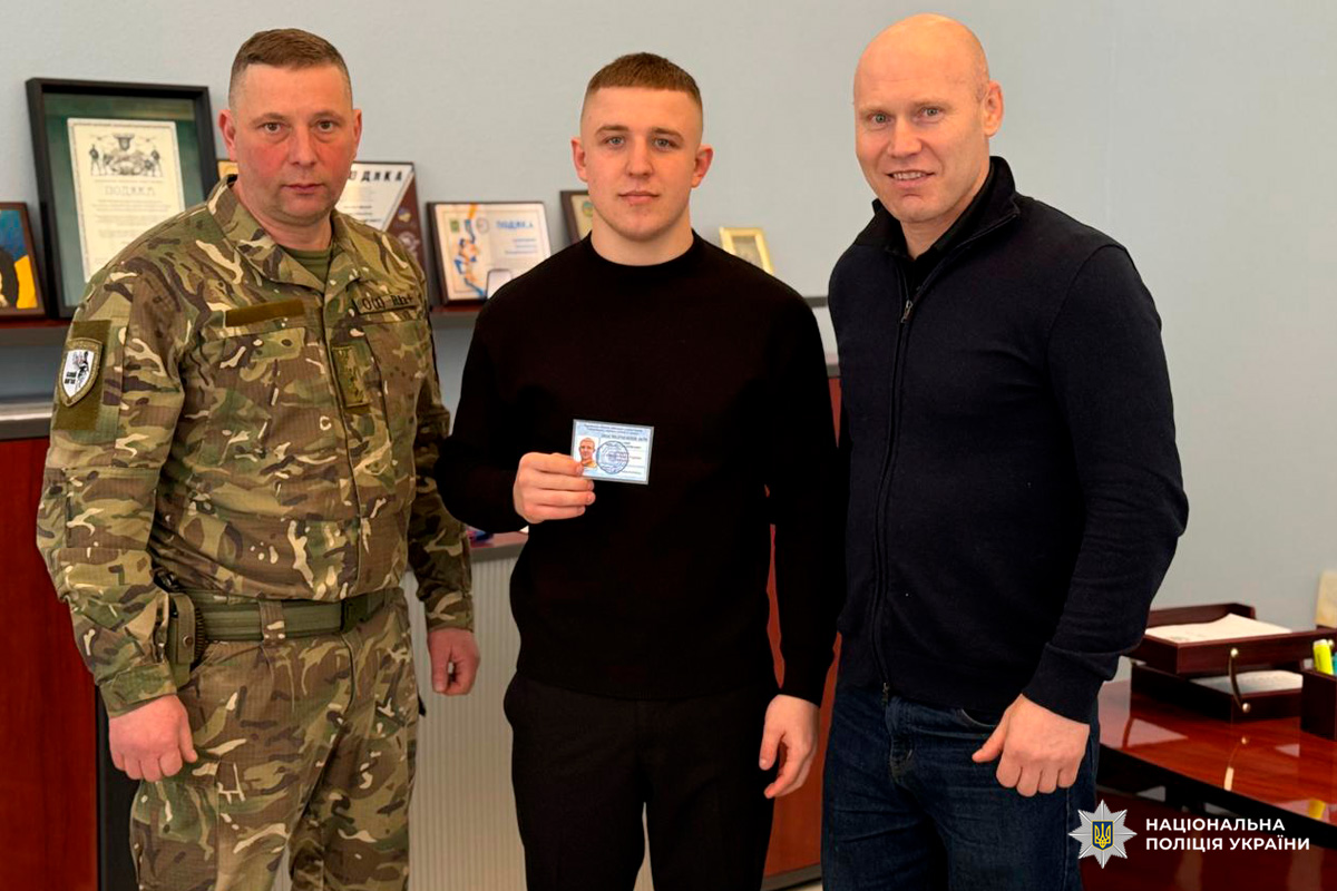 У приміщенні сфотографовано трьох людей: один у військовій формі, інший у цивільному одязі, а центральна особа тримає документ.