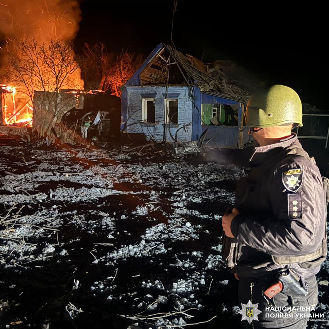Поліцейський у формі Національної поліції України фіксує наслідки пожежі: зруйнований будинок із обваленим дахом та палаючою спорудою поруч.