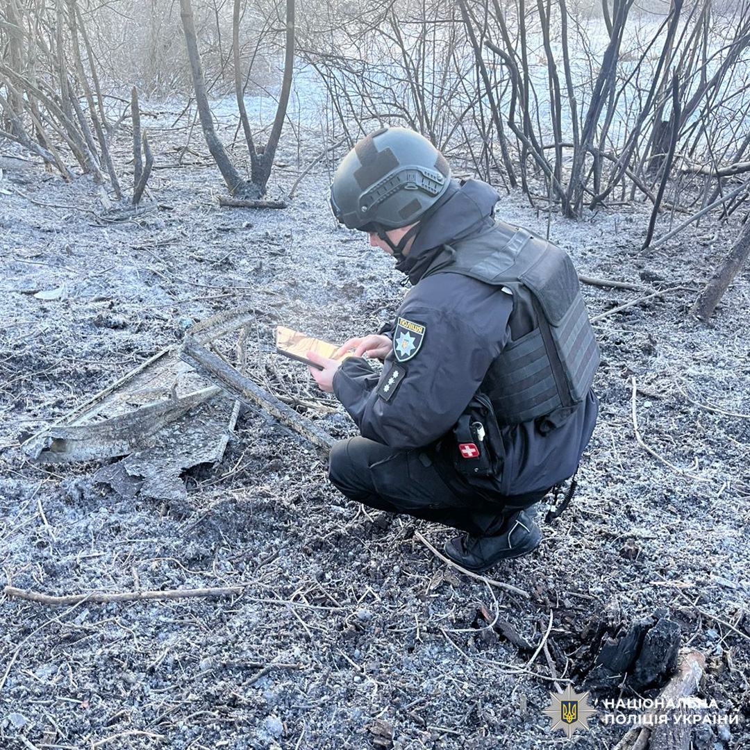 Поліцейський у бронежилеті та касці проводить огляд території з обгорілим ґрунтом і уламками.