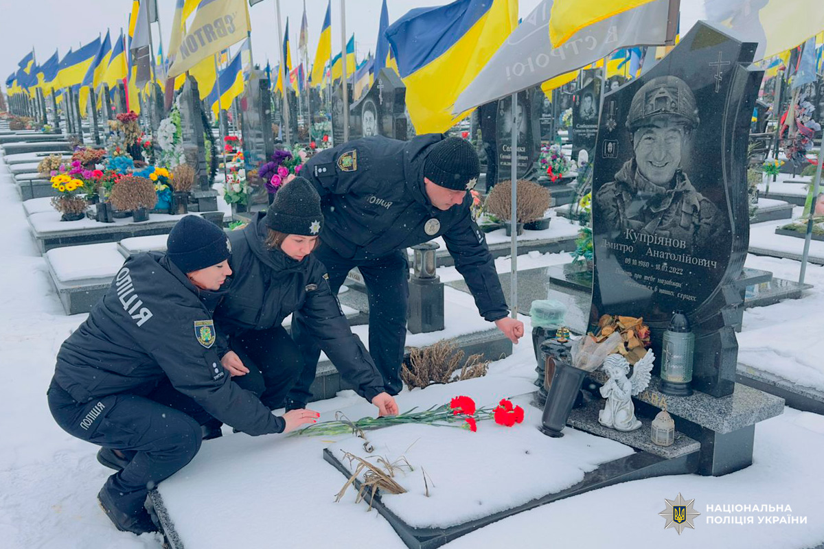Поліцейські в зимовій формі схиляються на колінах біля могили Дмитра Купріянова, покладаючи червоні квіти. Засніжене кладовище прикрашене українськими прапорами та вінками.