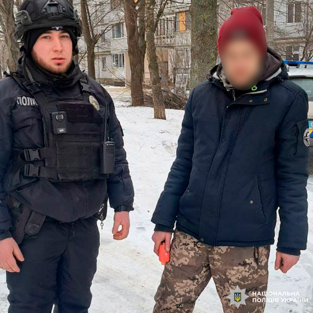 Поліцейський у бронежилеті та шоломі спілкується з чоловіком у зимовому одязі, поруч видно службовий автомобіль.