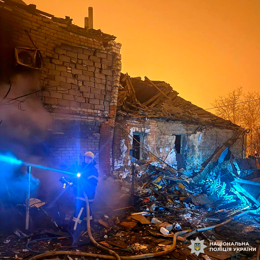 Пожежник гасить вогонь біля зруйнованої цегляної будівлі з обваленим дахом, навколо уламки та руїни.