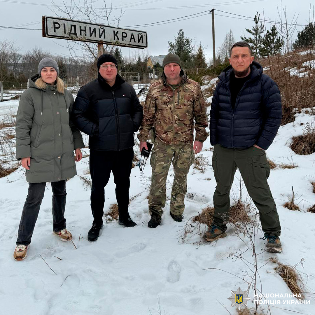 Чотири особи у зимовому одязі стоять на снігу біля таблички “Рідний край”.