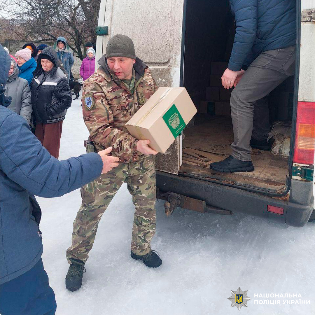 Працівник у камуфляжній формі передає коробку з гуманітарною допомогою іншій людині біля автомобіля. У кадрі видно чергу людей у зимовому одязі.