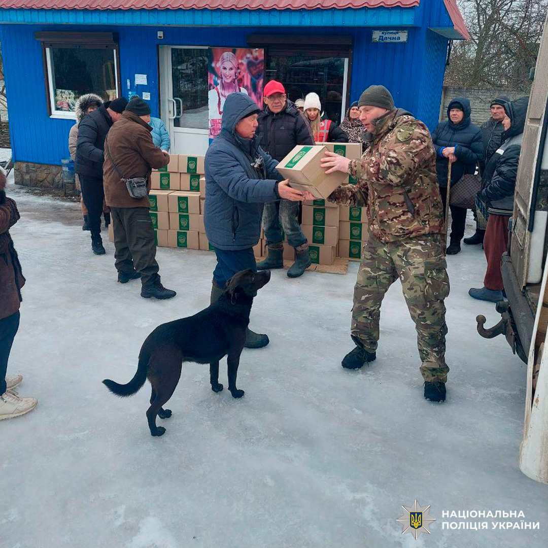 Поліцейський у камуфляжі передає коробку гуманітарної допомоги чоловікові у зимовому одязі біля будівлі з табличкою “вулиця Дачна”. На передньому плані чорний собака, у фоні  люди та складені коробки.