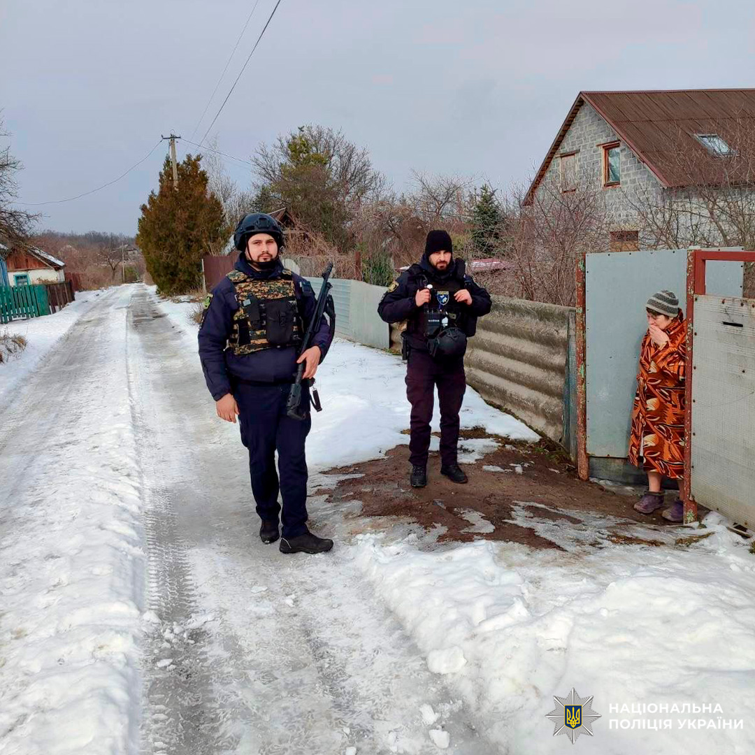 Двоє поліцейських у зимовій формі та спорядженні виконують службові обов’язки на засніженій вулиці приватного сектору. Поруч біля воріт стоїть літня жінка, закутана в теплу ковдру.