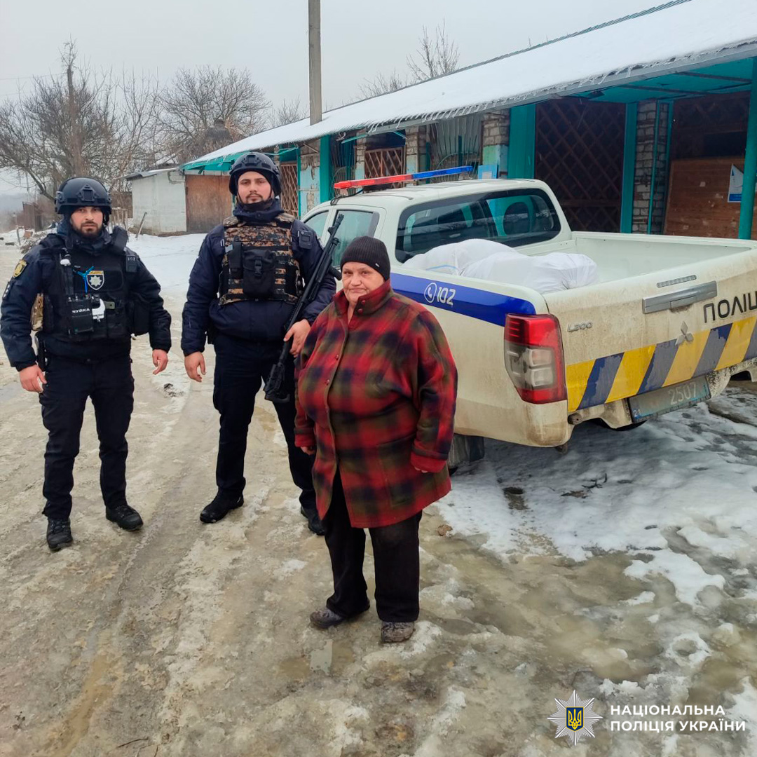 Двоє поліцейських у тактичному спорядженні стоять поруч із літньою жінкою біля службового автомобіля «Поліція» на засніженій дорозі.