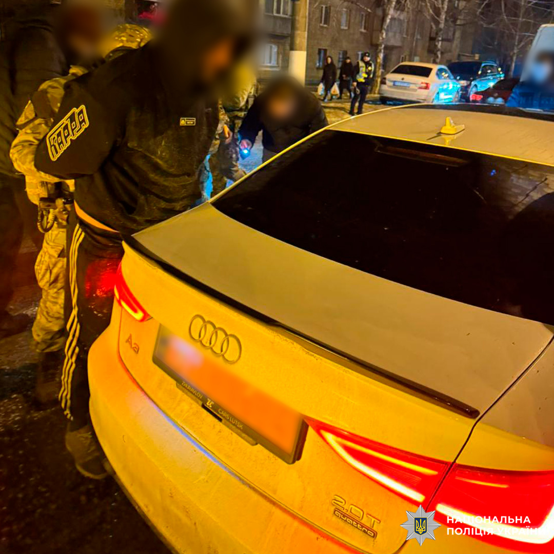 Поліцейські у тактичному спорядженні здійснюють обшук підозрюваного поруч із припаркованим автомобілем.