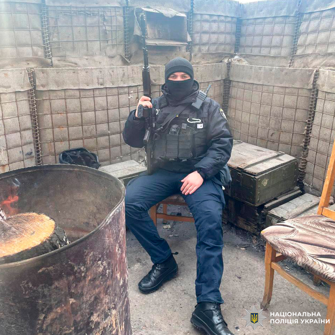 Особа у тактичному спорядженні та балаклаві сидить на стільці у фортифікованій зоні, тримаючи гвинтівку.