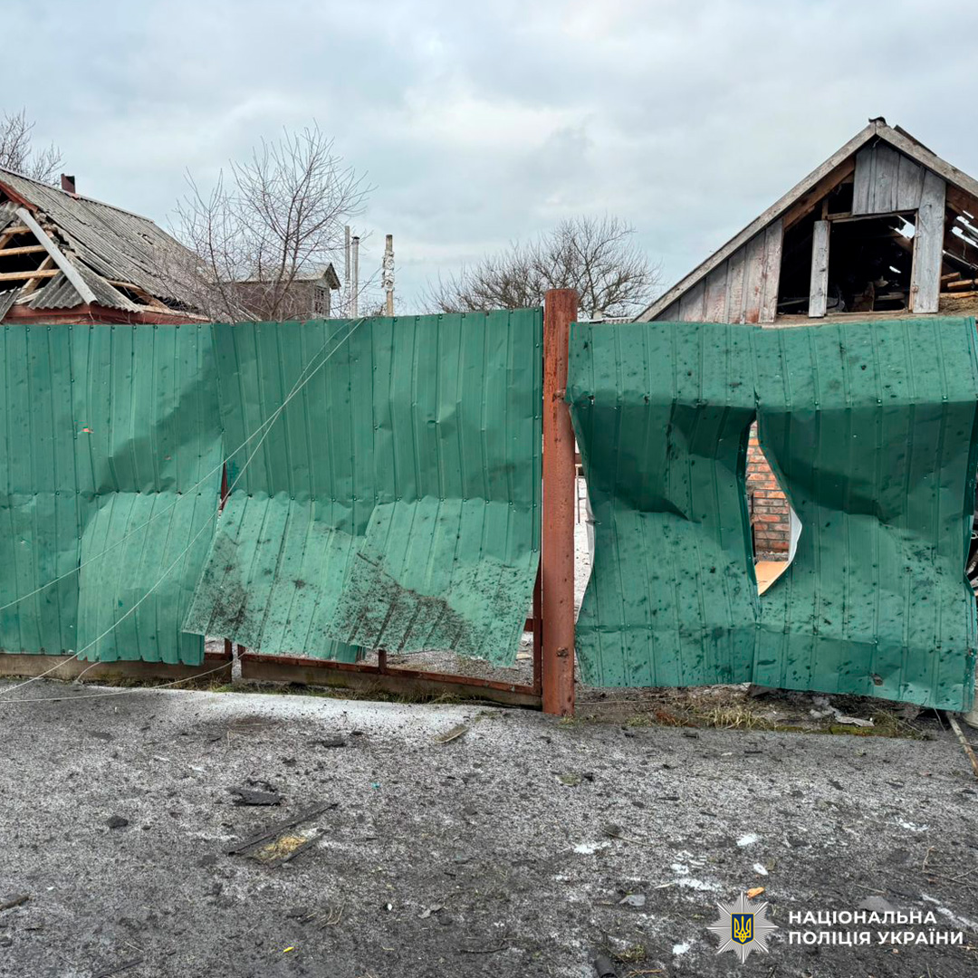 Пошкоджений металевий паркан із численними пробоїнами та деформаціями, за ним - будинки з вибитими дахами.