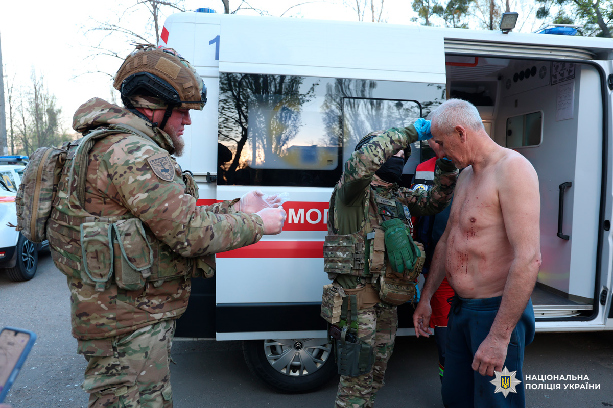 Поліцейські та військові надають медичну допомогу пораненому чоловікові біля карети швидкої допомоги.