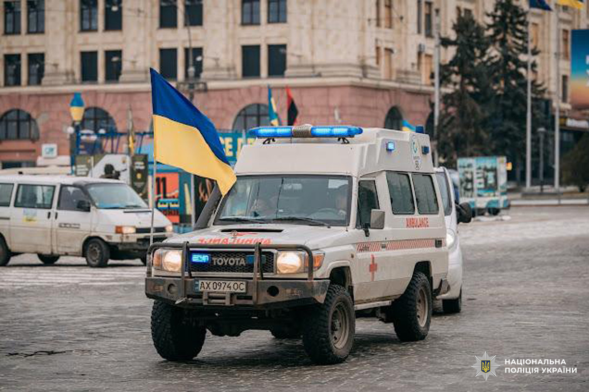 Службовий автомобіль швидкої допомоги з українським прапором прямує центральною вулицею, на задньому плані видно інші машини та будівлю з арочними вікнами