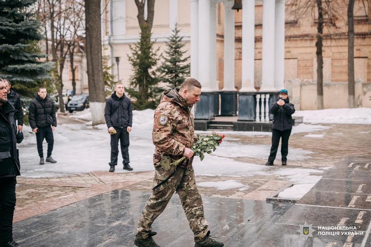 Чоловік у військовій формі кладе квіти до чорного пам’ятного каменя з іменами полеглих. На тлі  люди у темному одязі та колонада