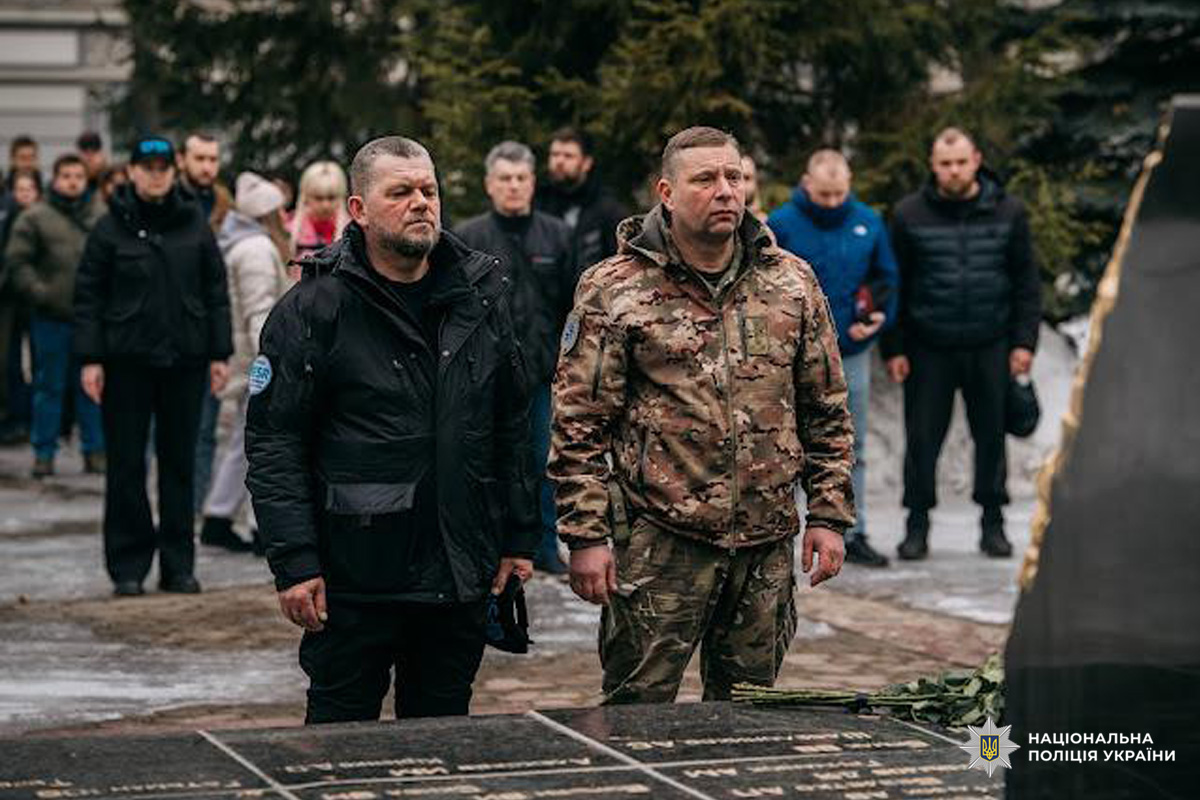 Учасники офіційного заходу стоять біля меморіалу з квітами. Присутні представники поліції.