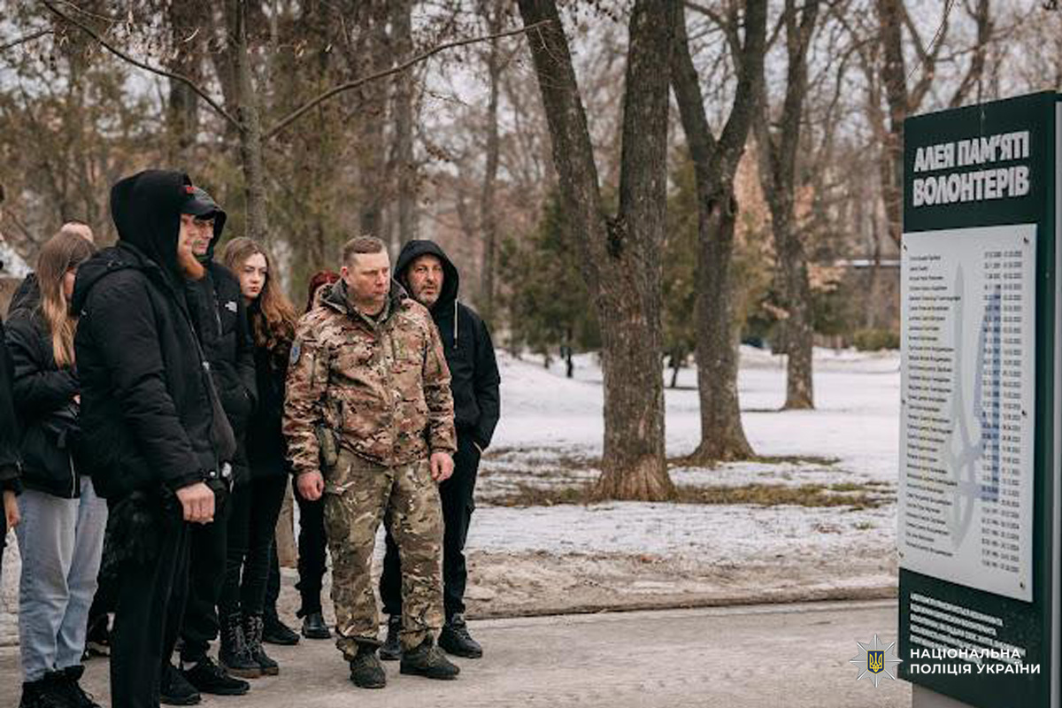 Група людей у зимовому парку стоїть біля пам’ятного знаку “Алея пам’яті волонтерів”, на якому викарбувані імена та дати
