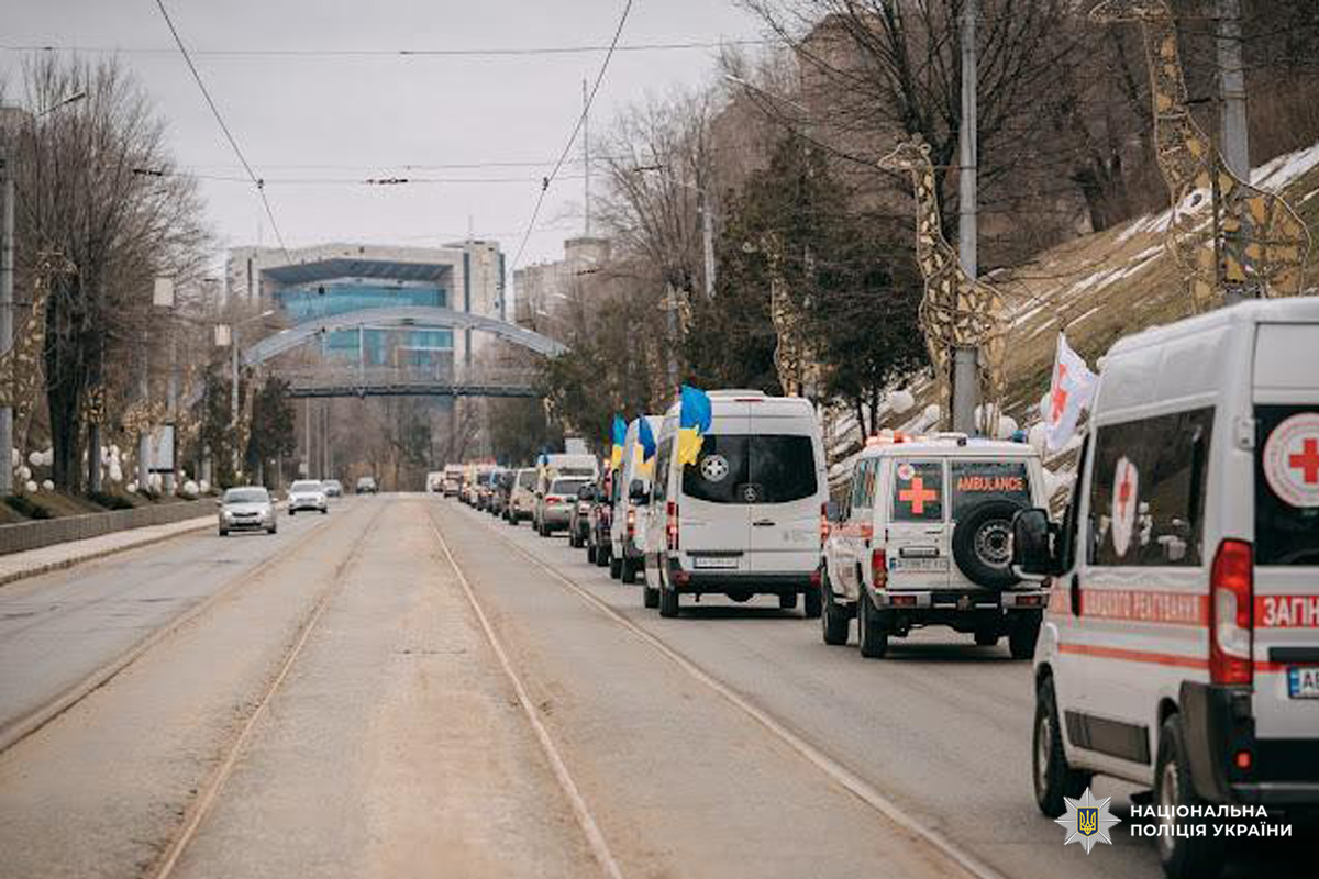 Колона автомобілів швидкої допомоги та екстрених служб із прапорами України та символами Червоного Хреста рухається міською вулицею. На тлі — сучасна будівля зі скляним фасадом та арочний пішохідний міст