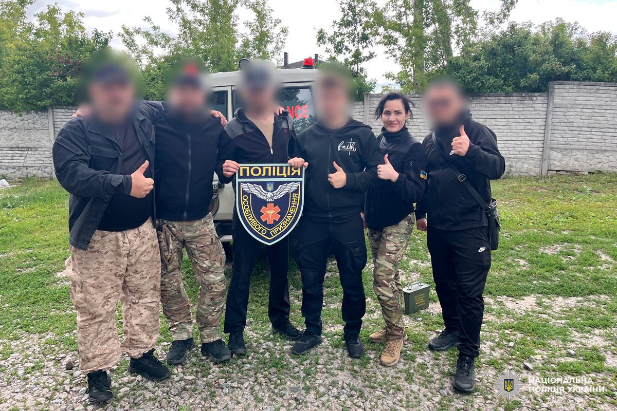 Група співробітників у тактичному спорядженні позує біля автомобіля швидкої допомоги, один тримає емблему спеціального підрозділу поліції.