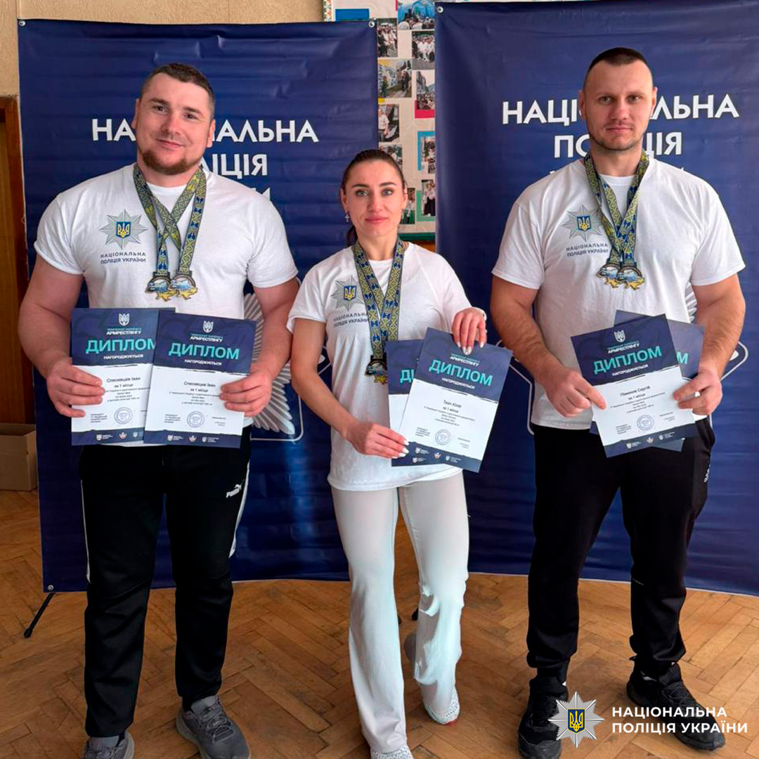Троє працівників Національної поліції України у білих футболках із емблемою, тримають дипломи та медалі на тлі синіх банерів.