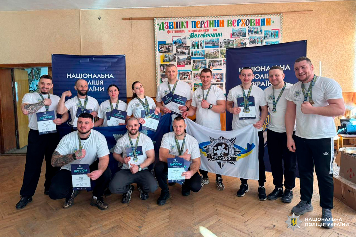 Група з дванадцяти представників Національної поліції України у білих футболках із медалями та дипломами, позують перед синіми банерами.