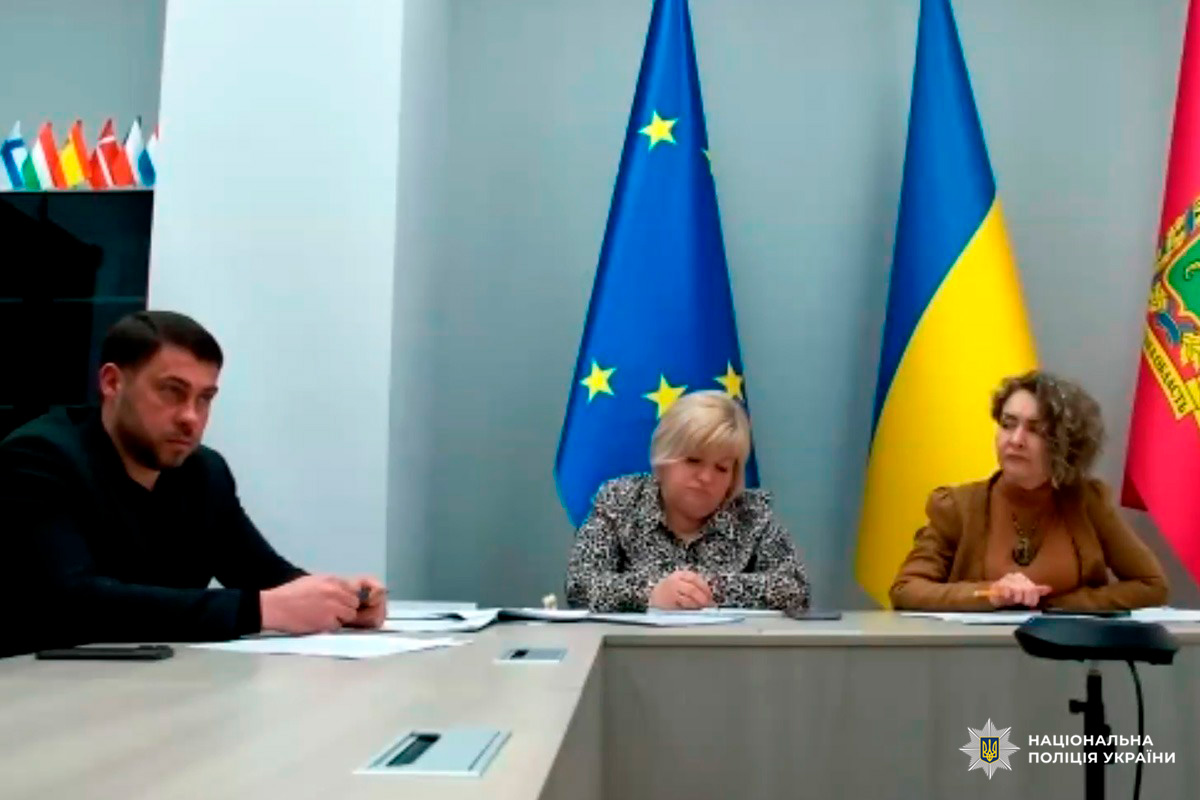 Троє учасників офіційної зустрічі сидять за столом, позаду прапори ЄС, України та регіональний прапор.