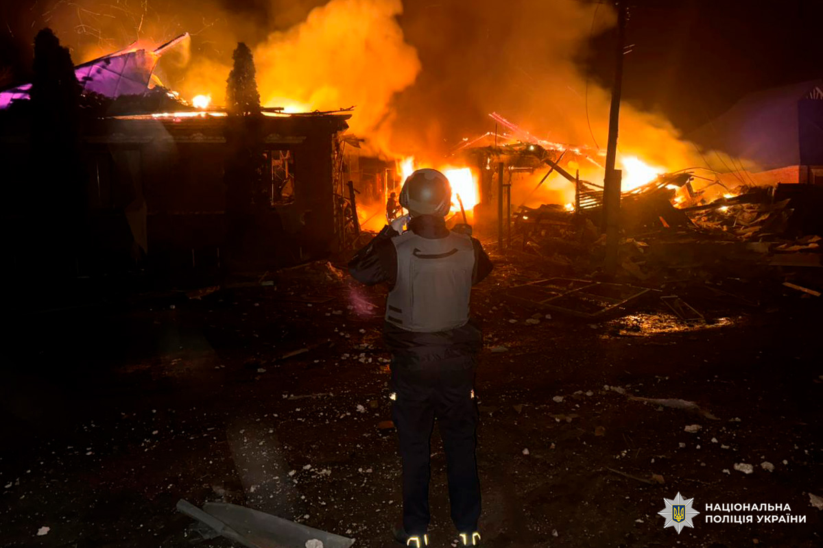 Полум’я знищує будівлю, а людина у спорядженні стоїть серед руїн.