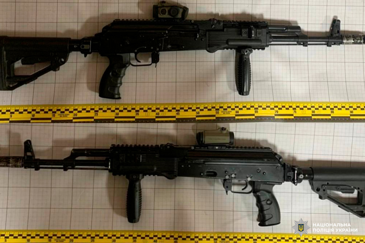 Два автомати з тактичним обвісом та коліматорними прицілами на фоні криміналістичної лінійки.