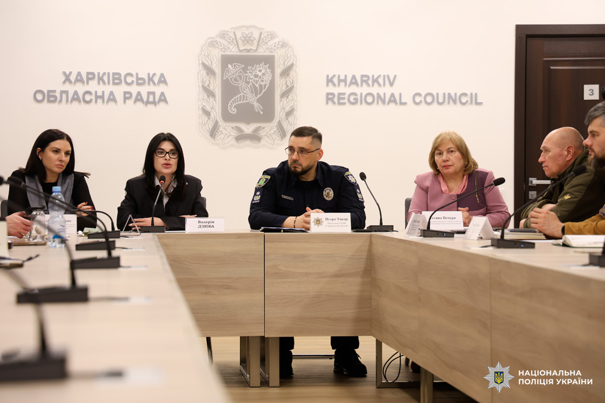 П'ятеро учасників офіційної зустрічі сидять за великим дерев'яним столом з мікрофонами та табличками. У центрі - чоловік у формі поліції, поруч жінки та чоловіки у діловому та мілітарі одязі.