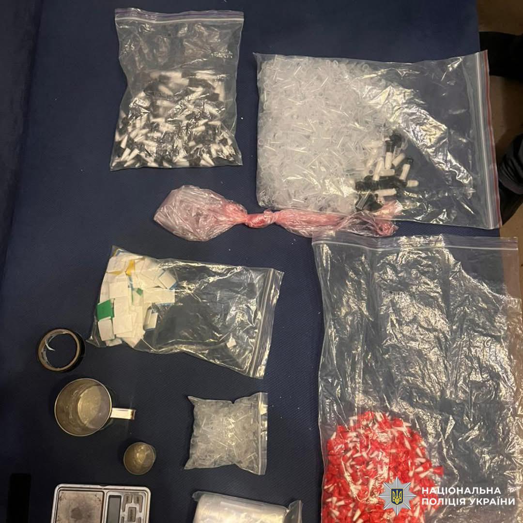 Пакети з капсулами, контейнери, ваги та інші предмети, розкладені на темній поверхні.