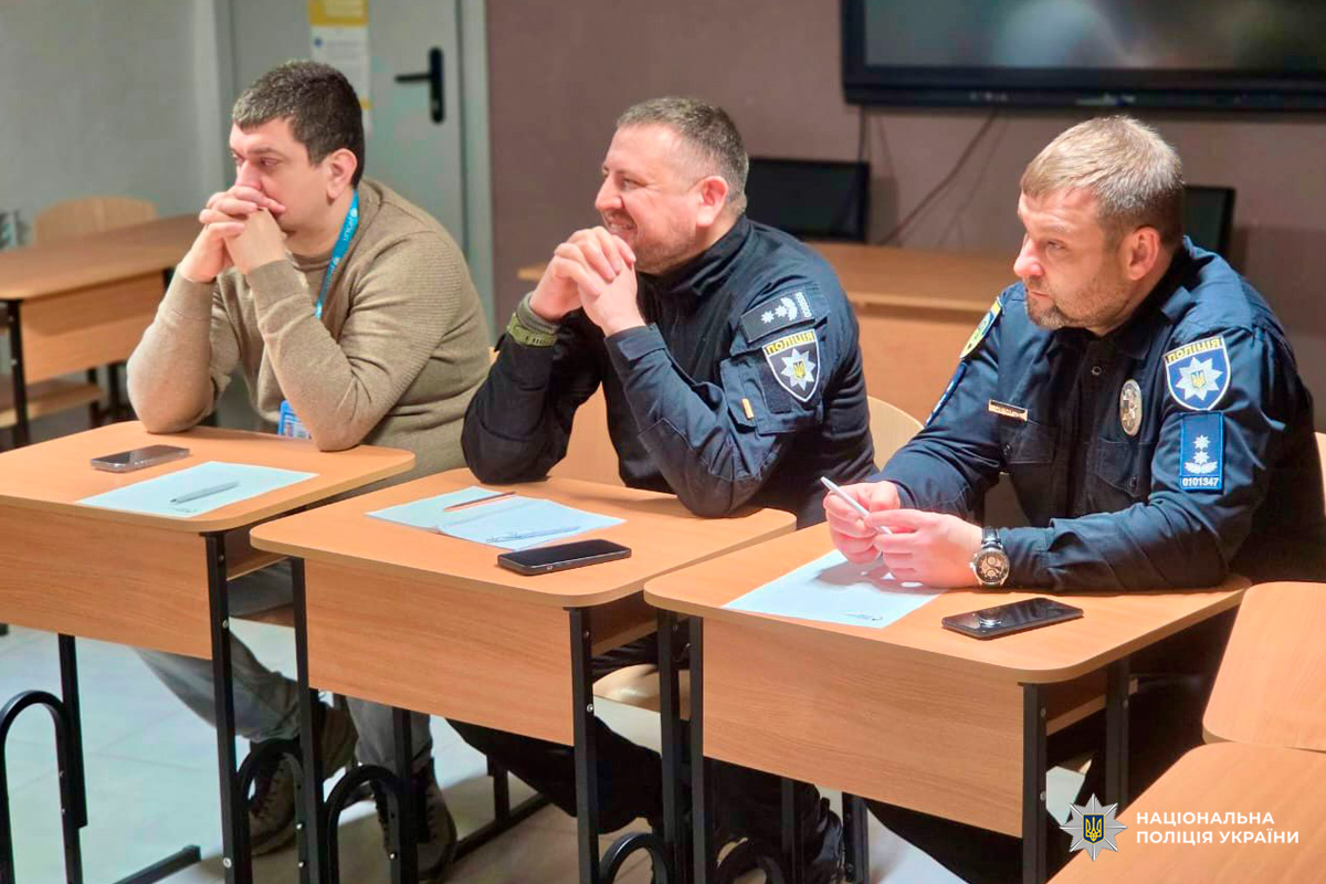 Двоє поліцейських у формі та цивільний учасник сидять за столами з документами й телефонами,