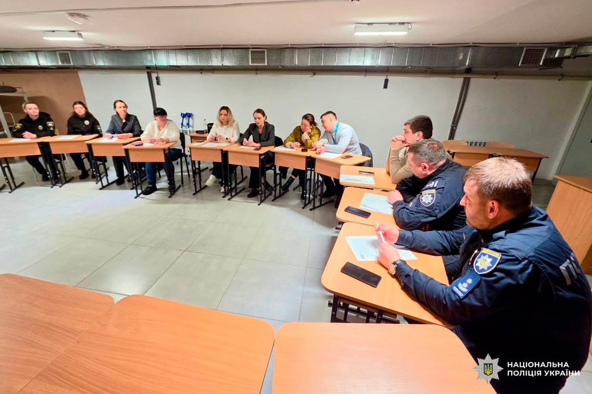 Учасники наради сидять за U‑подібними столами, серед них поліцейські у формі та цивільні особи,